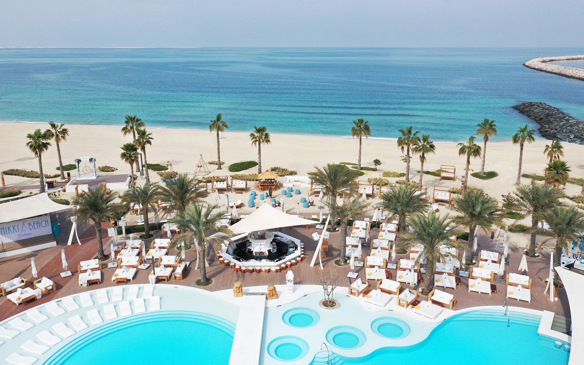 Nikki Beach Dubai seen from above: golden sand and turquoise sea lined with palm trees, white daybeds and umbrellas